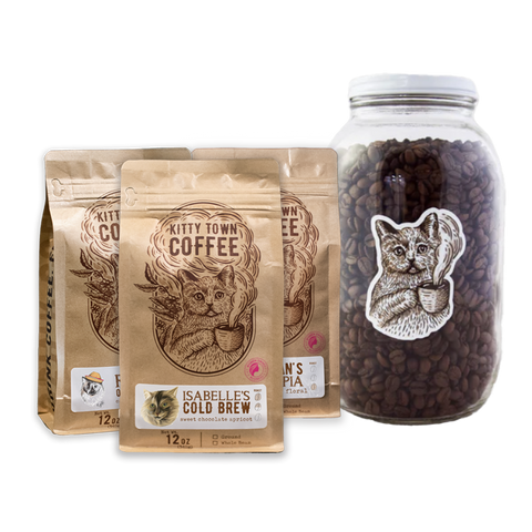 A collection of coffee-related items including three bags of coffee beans labeled 'Kitty Town Coffee' with different names, and a large 64oz glass jar with a metal lid, containing coffee beans and featuring a cat graphic.