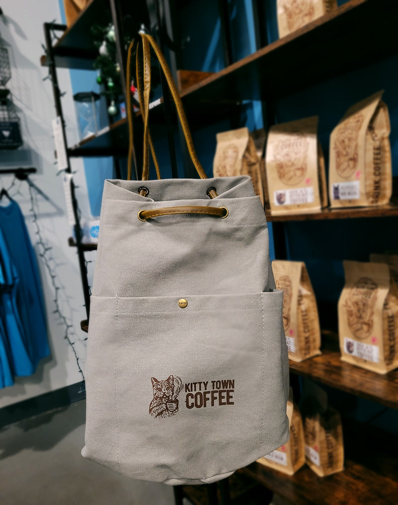 A gray bucket style backpack with the Kitty Town Coffee logo on the front, with a background of a shop behind it