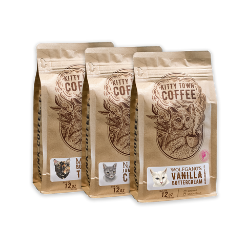 Three 12 oz bags of Kitty Town Coffee flavored coffee, with Wolfgang's Vanilla Buttercream in front and Mika's and Nadia's bags behind, each labeled with a photo of its cat