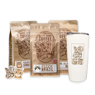 A gift set of Kitty Town Coffee products, including three bags of coffee (Fezzik, The Albert Blend, and Simba's Sumatra), a white mug with a logo, and two stickers with cat graphics.